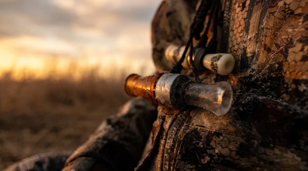 Duck calls hanging from a lanyard against a hunter's camouflage jacket at sunset