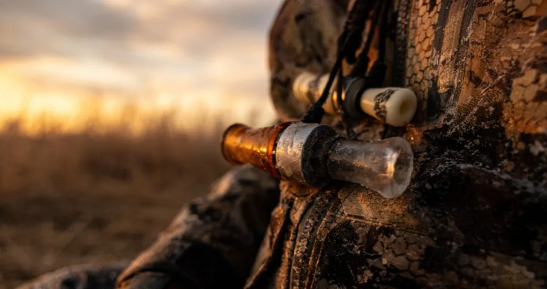 Duck calls hanging from a lanyard against a hunter's camouflage jacket at sunset