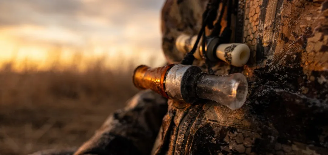 Duck calls hanging from a lanyard against a hunter's camouflage jacket at sunset